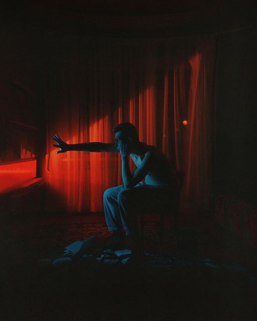 A man sitting in a dimly lit room reaches toward a dramatic red light, creating a mysterious ambiance.