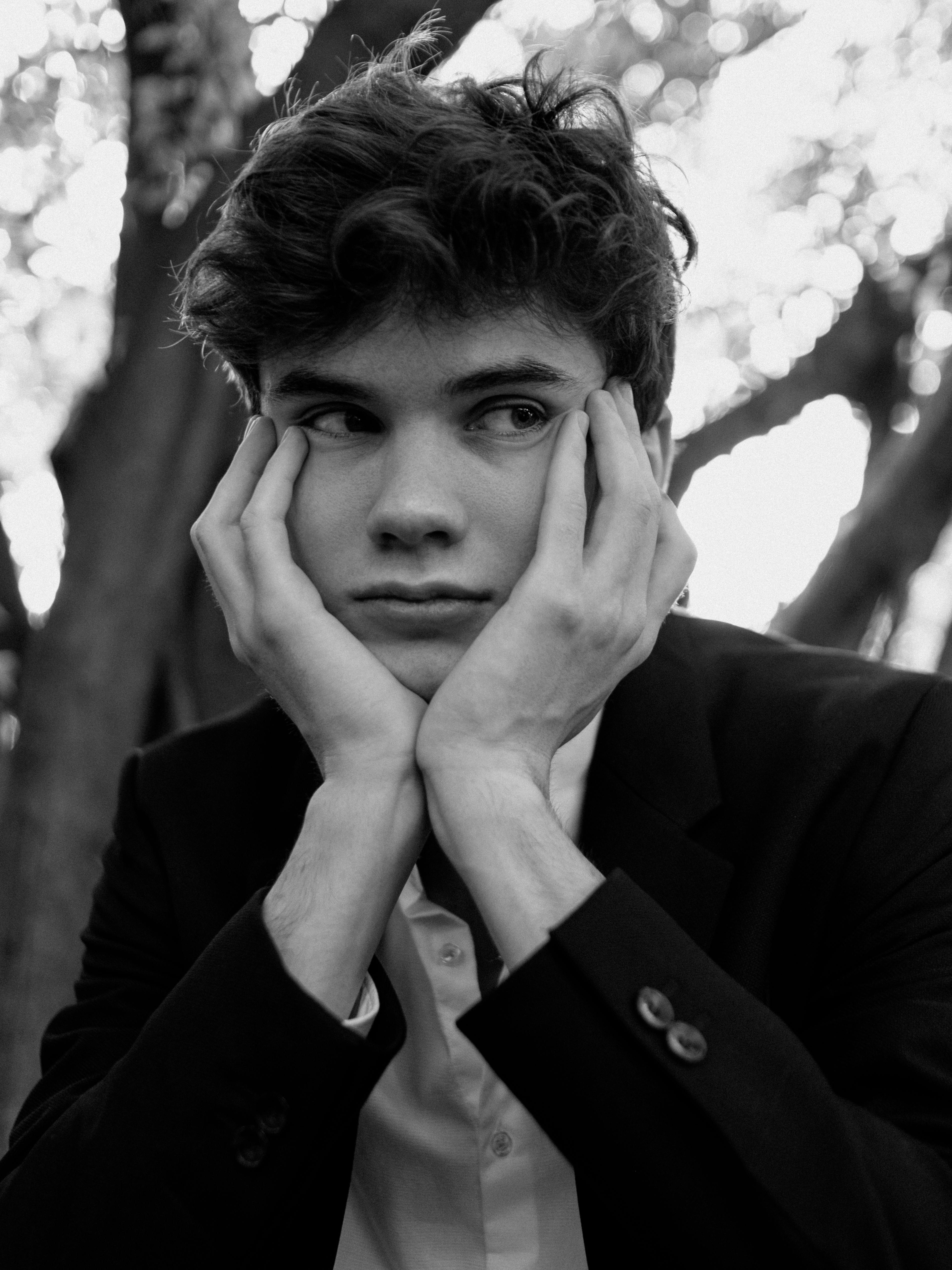 Black and white portrait of a young man with curly hair in a suit resting his face in his hands outdoors.