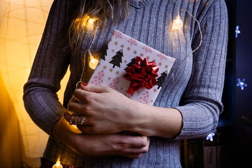 Free A woman holds a Christmas gift wrapped with a red bow and festive lights inside a cozy indoor setting. Stock Photo