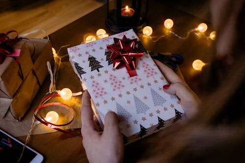 Person Holding Gift Box With Red Ribbon Beside String Lights