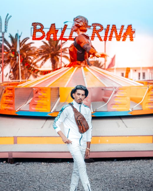Stylish man in white tracksuit poses at a colorful carnival backdrop with Ballerina ride.