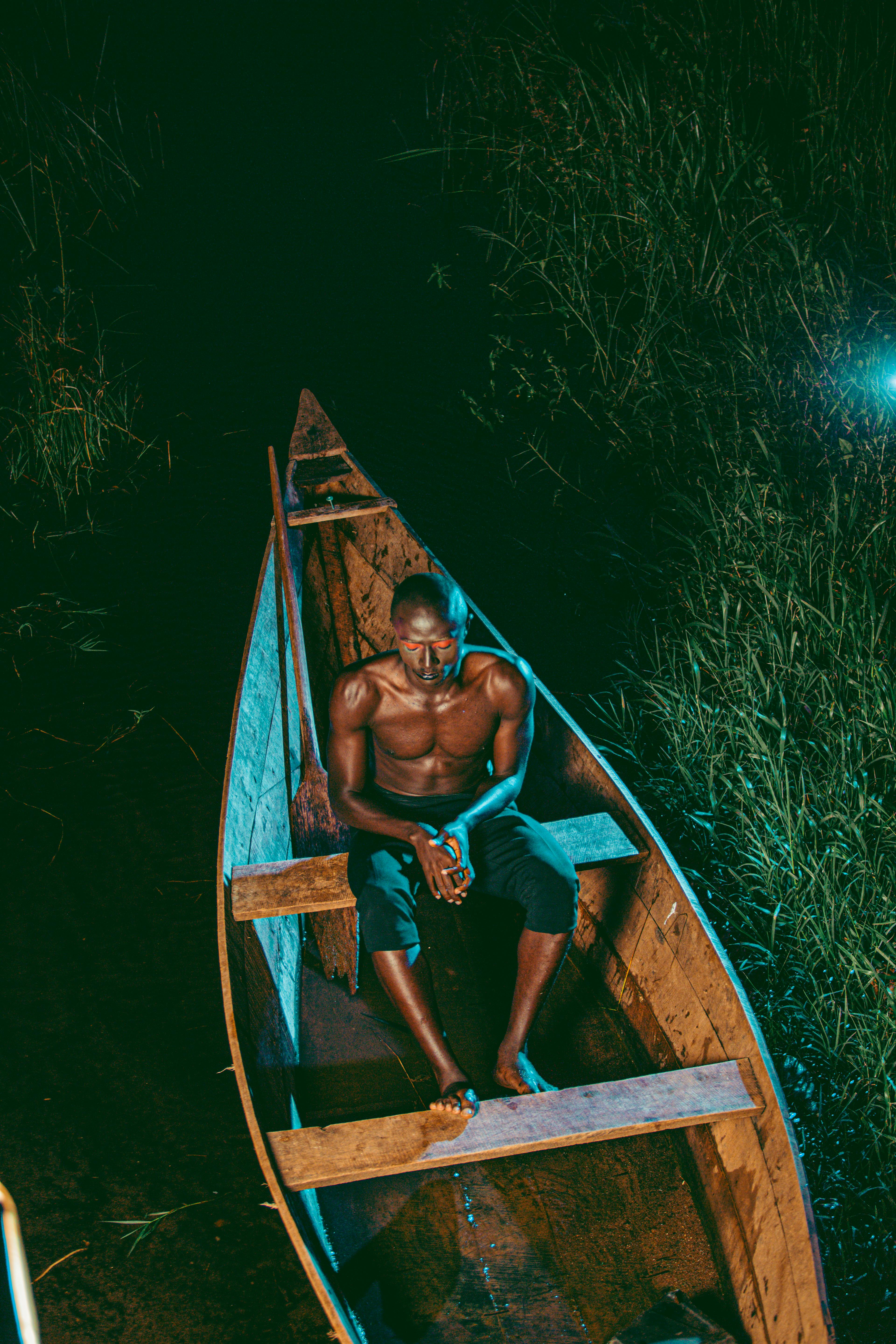 A shirtless man sits in a canoe amidst a nighttime wetland scene, exuding serenity.