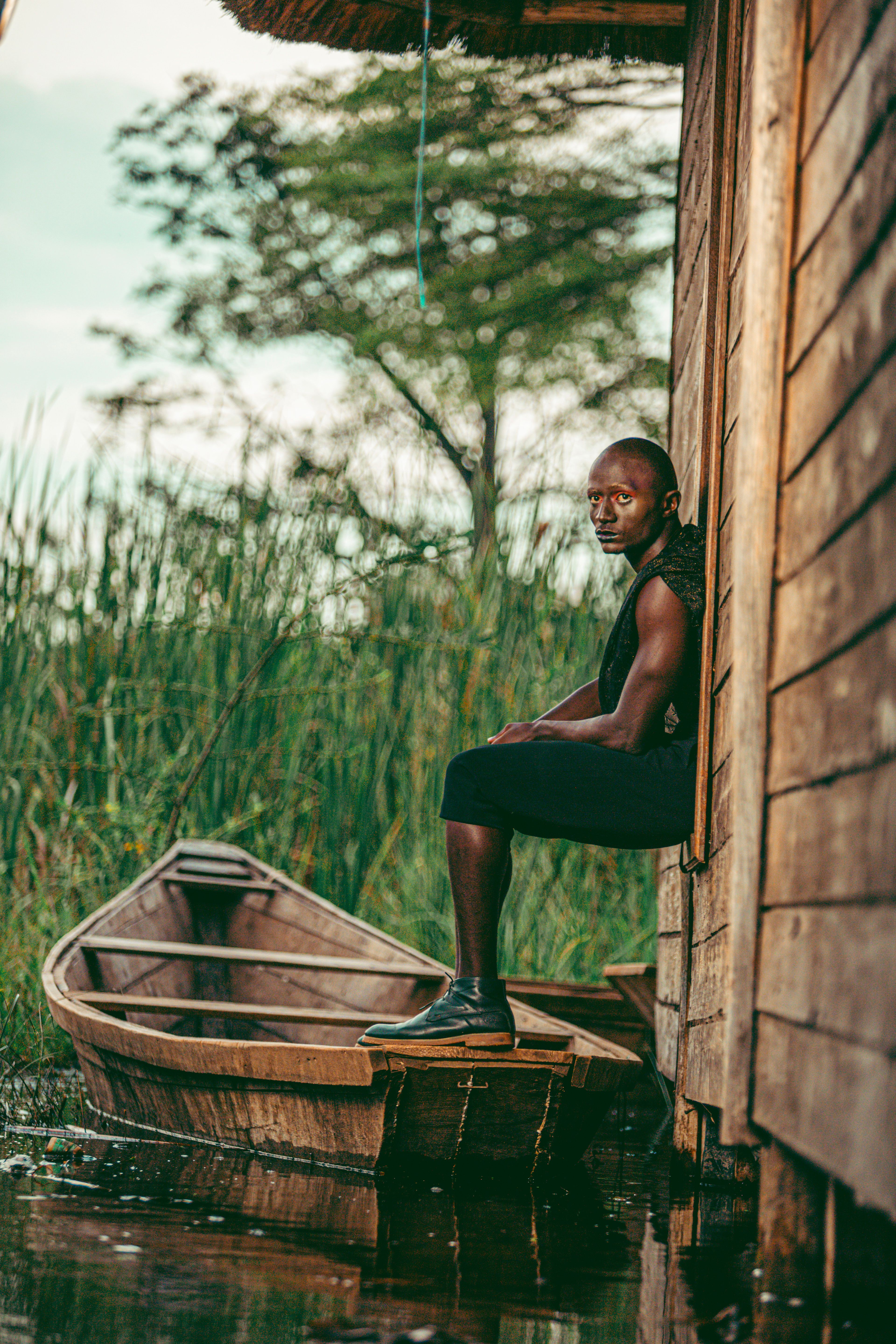 A man sits on a lakeside rustic wooden house near a boat, showing tranquility.