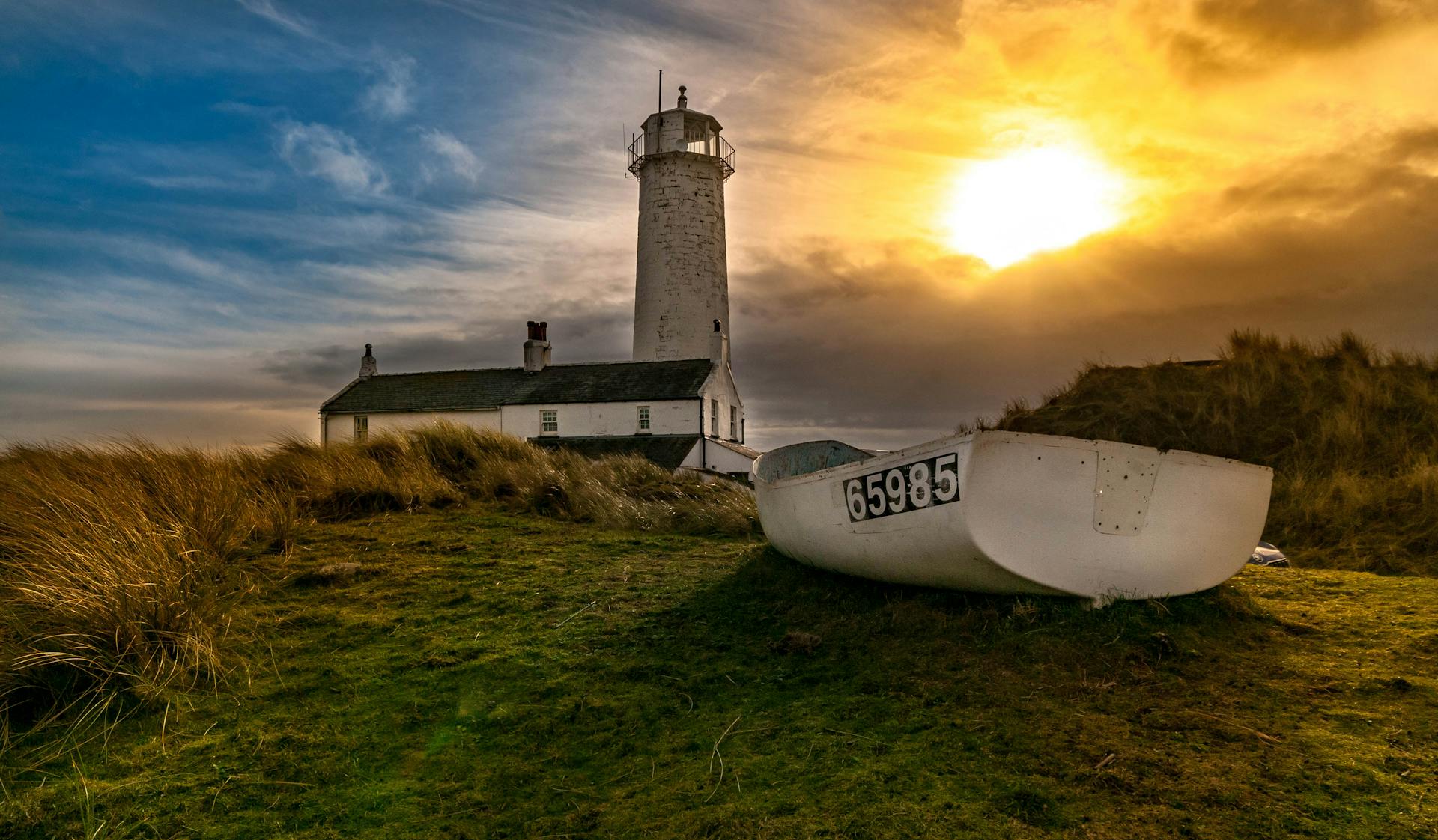 A breathtaking view of a lighthouse and boat at sunset with dramatic skies and coastal grasses.