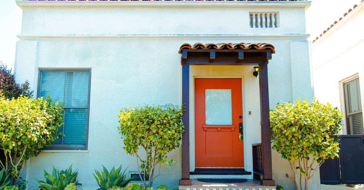 Vibrant Mediterranean-style home facade featuring an orange door, lush greenery, and classic architecture.