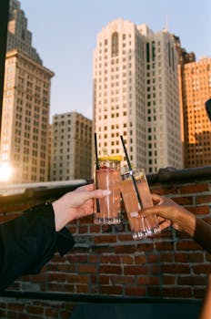 Two hands clinking cocktails on a rooftop with a city skyline in the background at sunset.