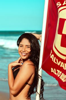 A woman enjoys a sunny day at the beach, smiling near a red safety flag.