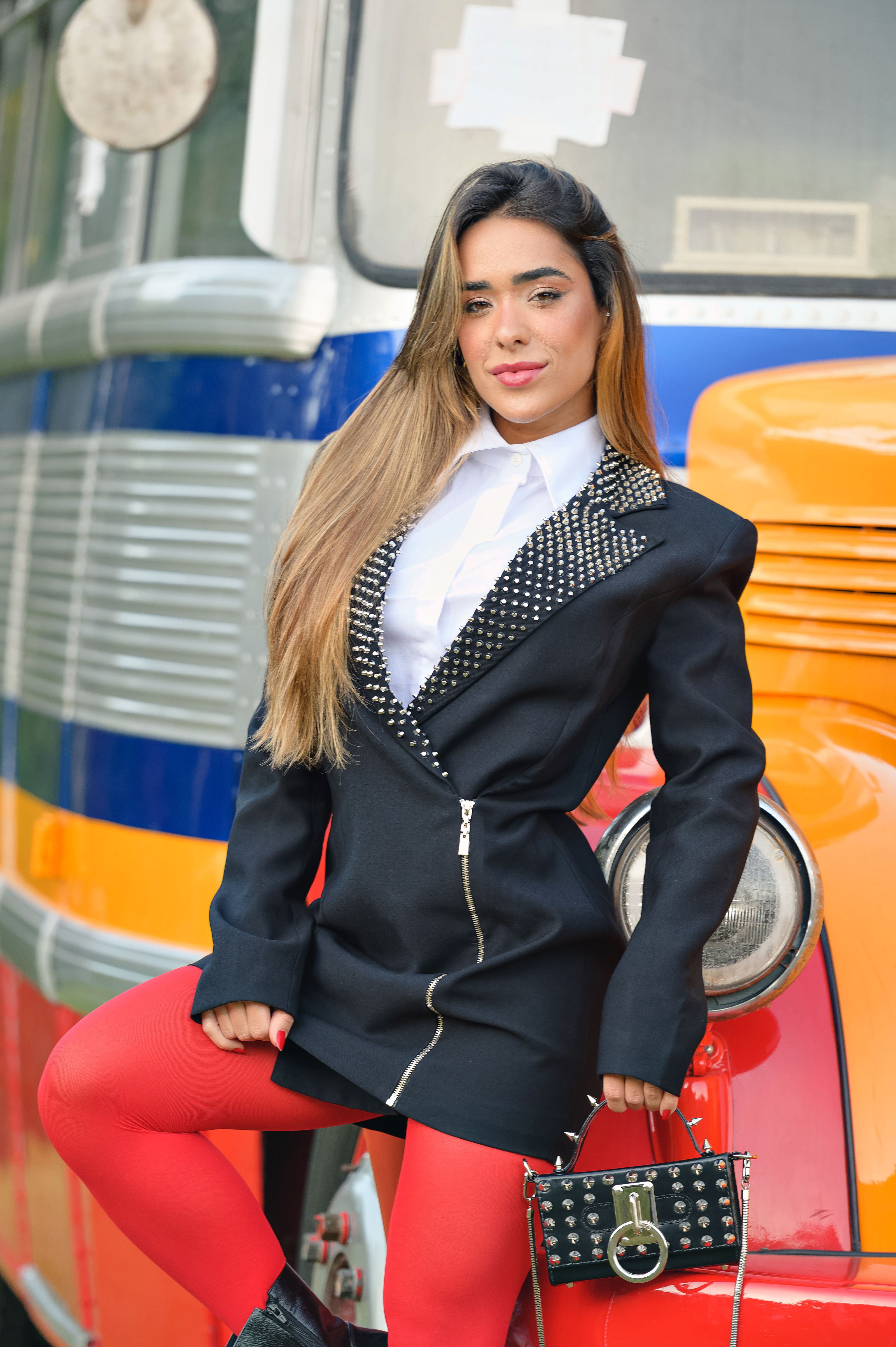 Mulher estilosa com blazer studded e meia-calça vermelha posando ao lado de um ônibus vintage.