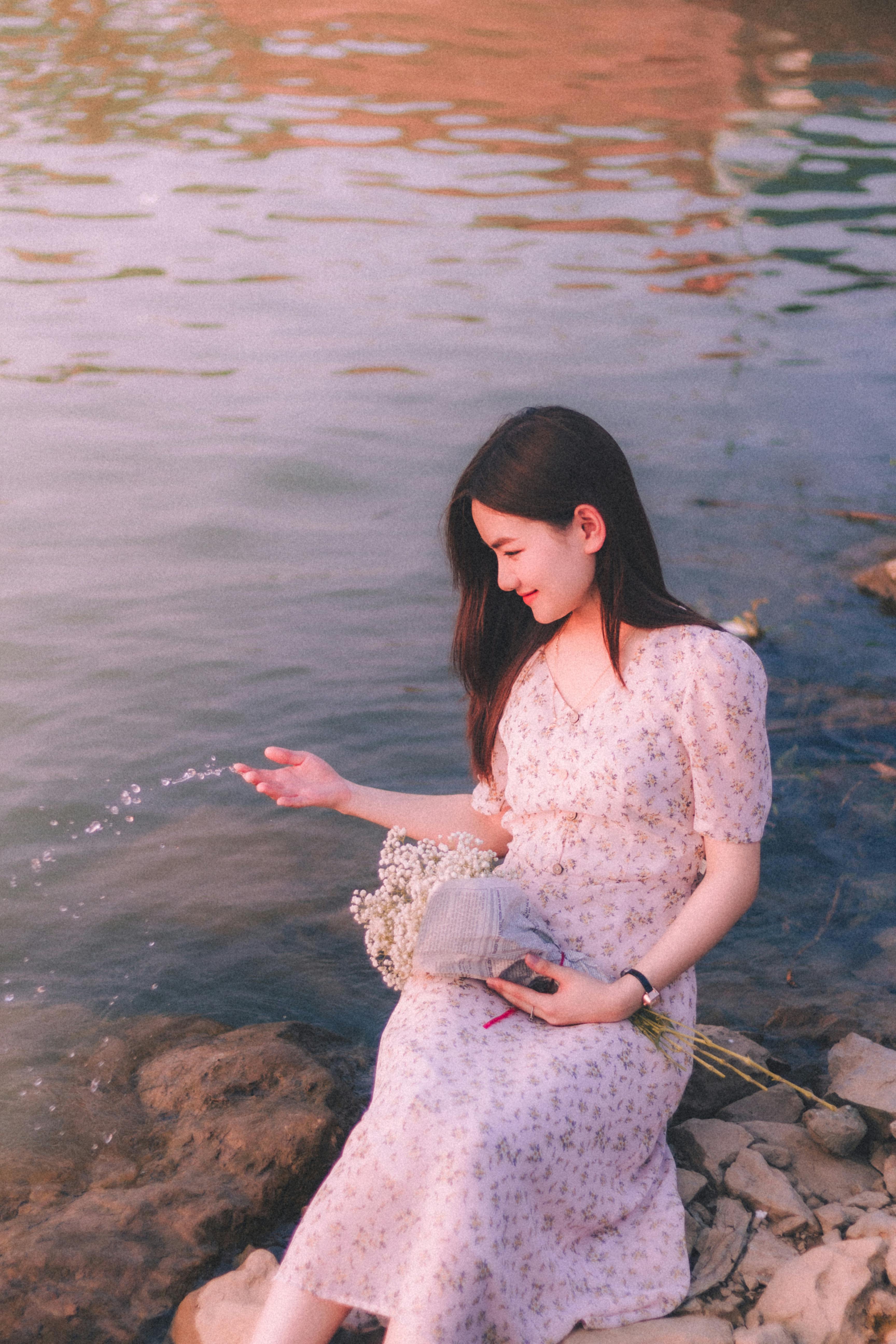 A young woman in a floral dress sits by the peaceful lakeside with a bouquet, enjoying a serene sunset.