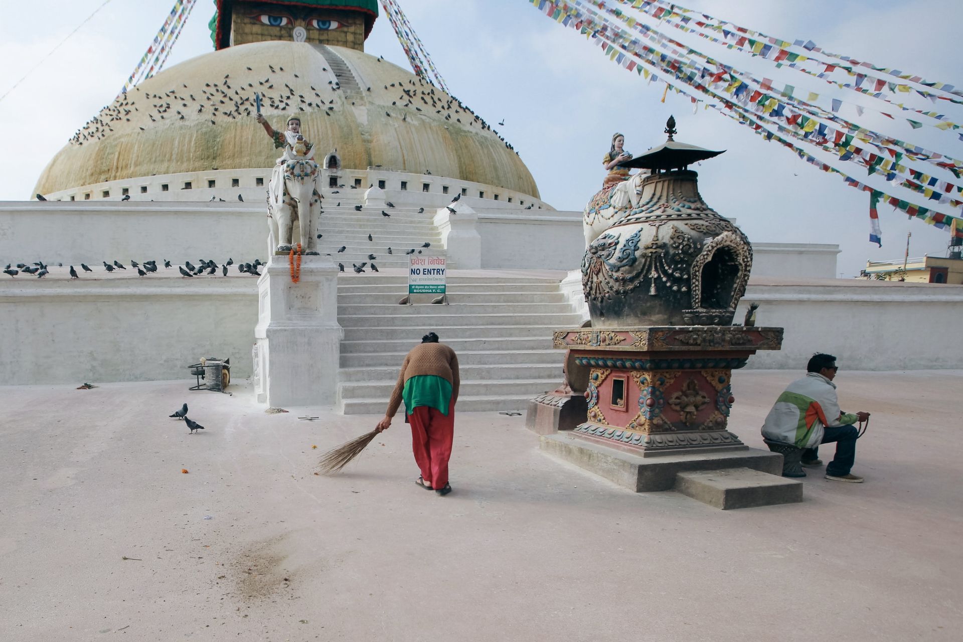 A local sweeps near Boudhanath Stupa in Kathmandu as pigeons gather on a sunny day.