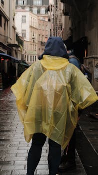 People walking on rainy city street wearing yellow raincoat. Urban scene with wet pavement and buildings.