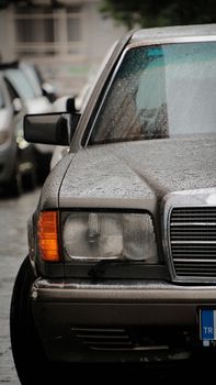 Close-up of a vintage car's headlight and hood in a rainy urban setting.