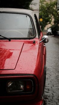 Close-up of a vintage red car parked on a rainy cobblestone city street.