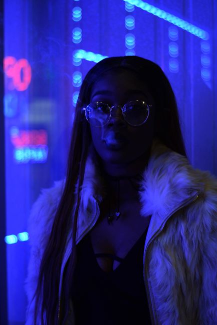 Stylish woman in a fur coat and glasses posing in a vibrant neon-lit city scene at night.