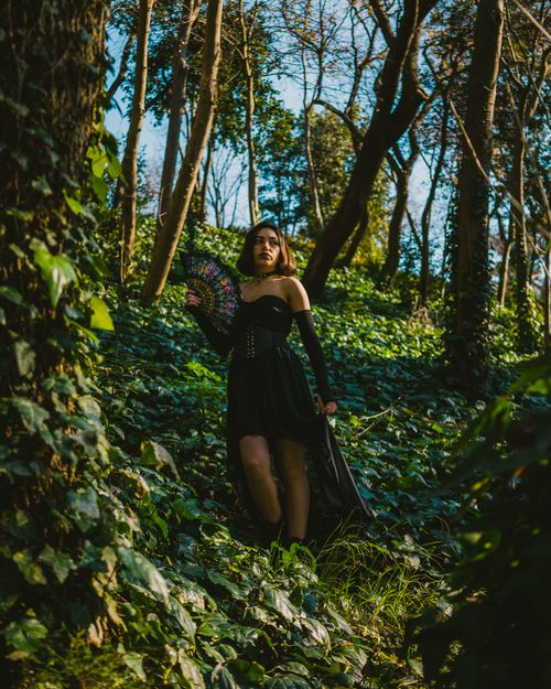 Free A stylish woman in a black dress posed in a lush green forest, holding a decorative fan. Stock Photo