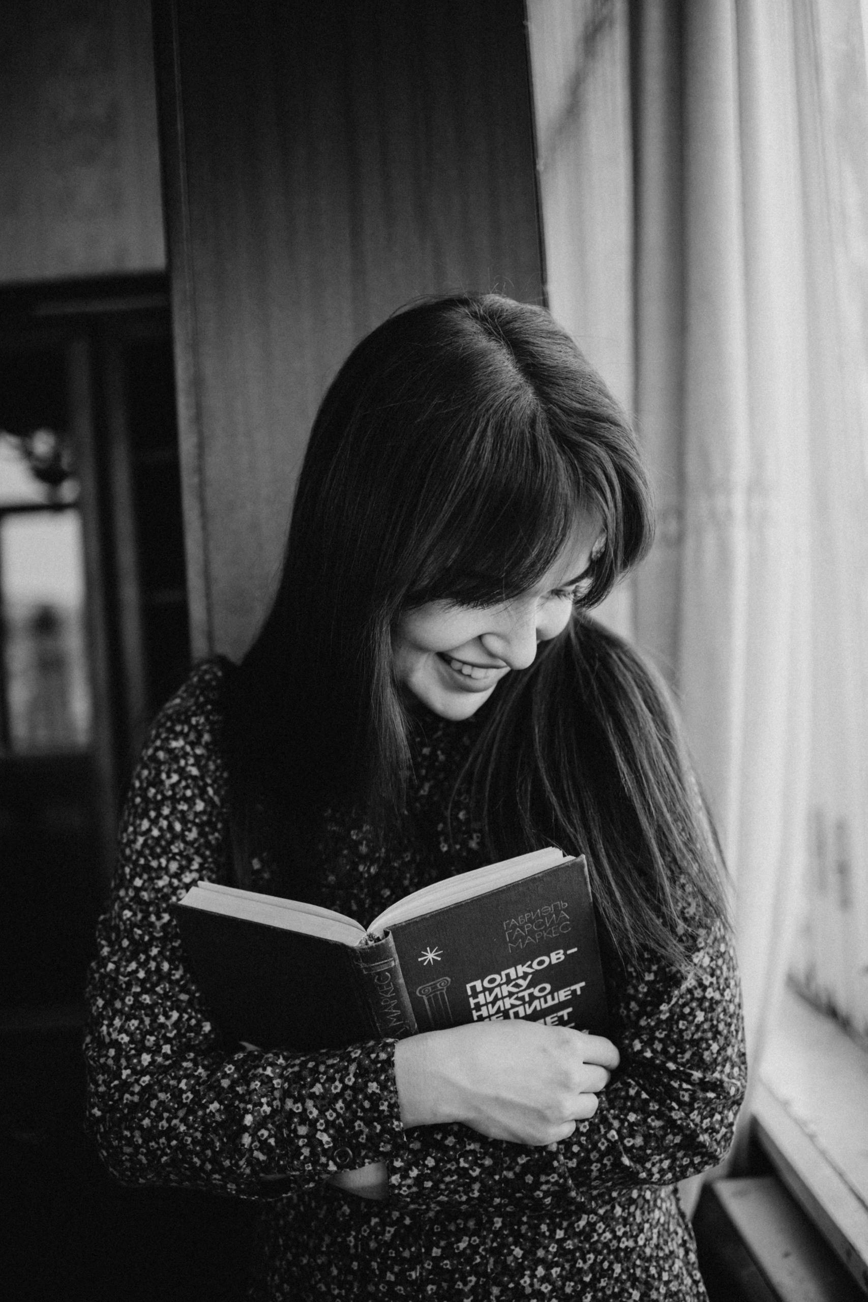 Black and white portrait of a smiling woman reading a book by the window, indoors, with a warm atmosphere.