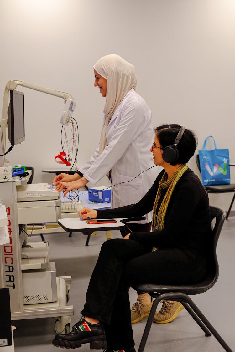 Woman With Headphones Sitting In An Examination Room And Woman In A Lab Coat Using A Computer 