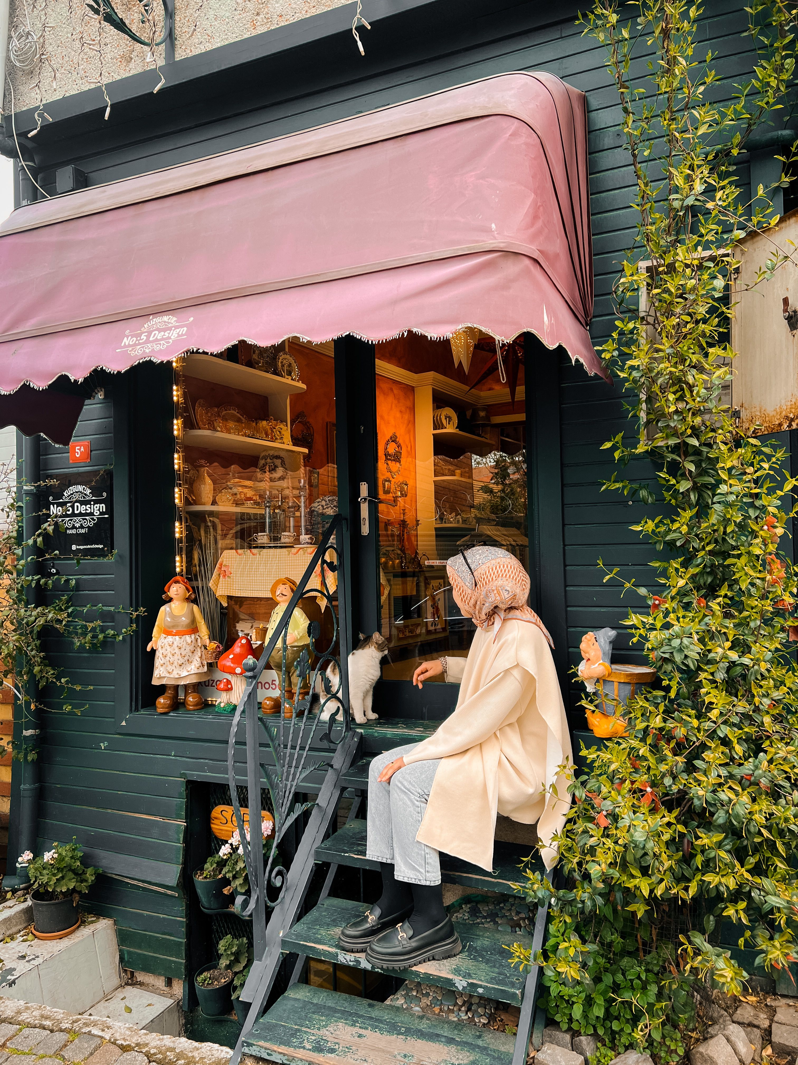 Woman in a hijab sits by a charming shop entrance with decorative elements.