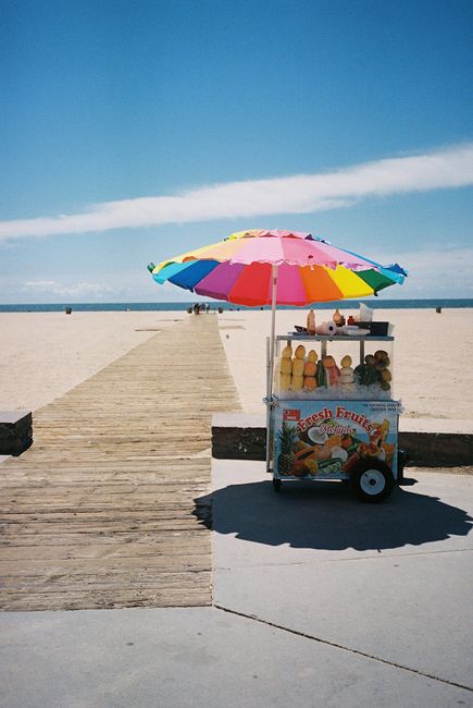Vibrant fruit stand with umbrella on sandy beach, perfect for summer relaxation.