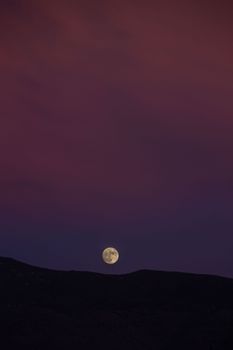 Stunning vertical shot of a full moon rising at dusk over dark hills with a colorful sky