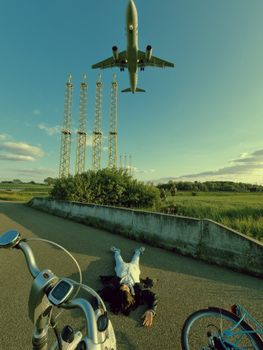 A person lies on a road beside bicycles as an airplane flies overhead in an outdoor setting.