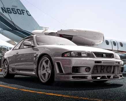 A silver sports car parked on tarmac with a private jet in the background in an urban setting.