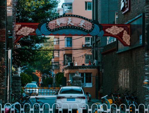 Free An urban street scene featuring a decorative gate, buildings, cars, and bicycles under a clear sky. Stock Photo