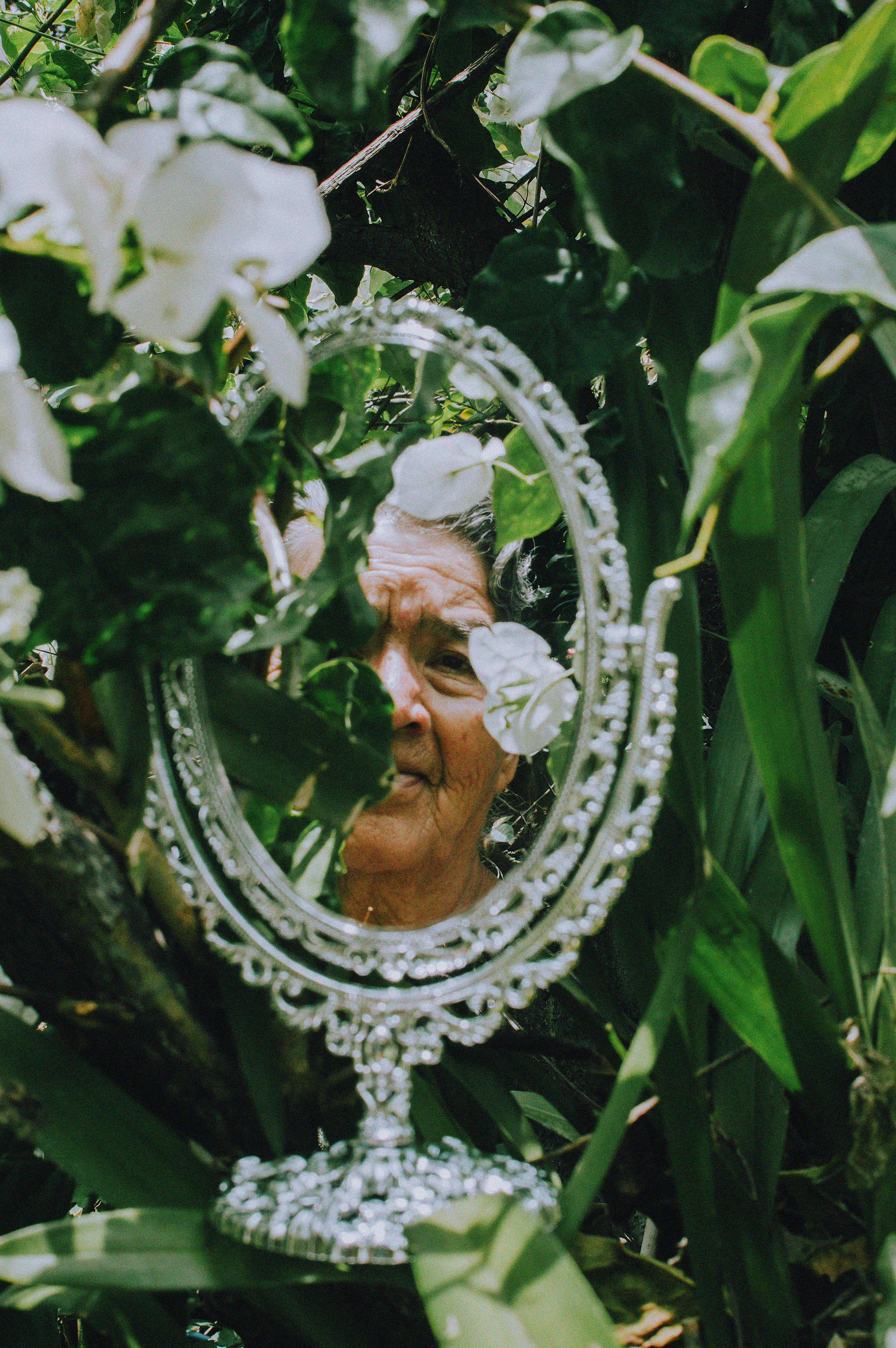 An elderly woman's face reflected in a mirror surrounded by lush green foliage and white flowers.