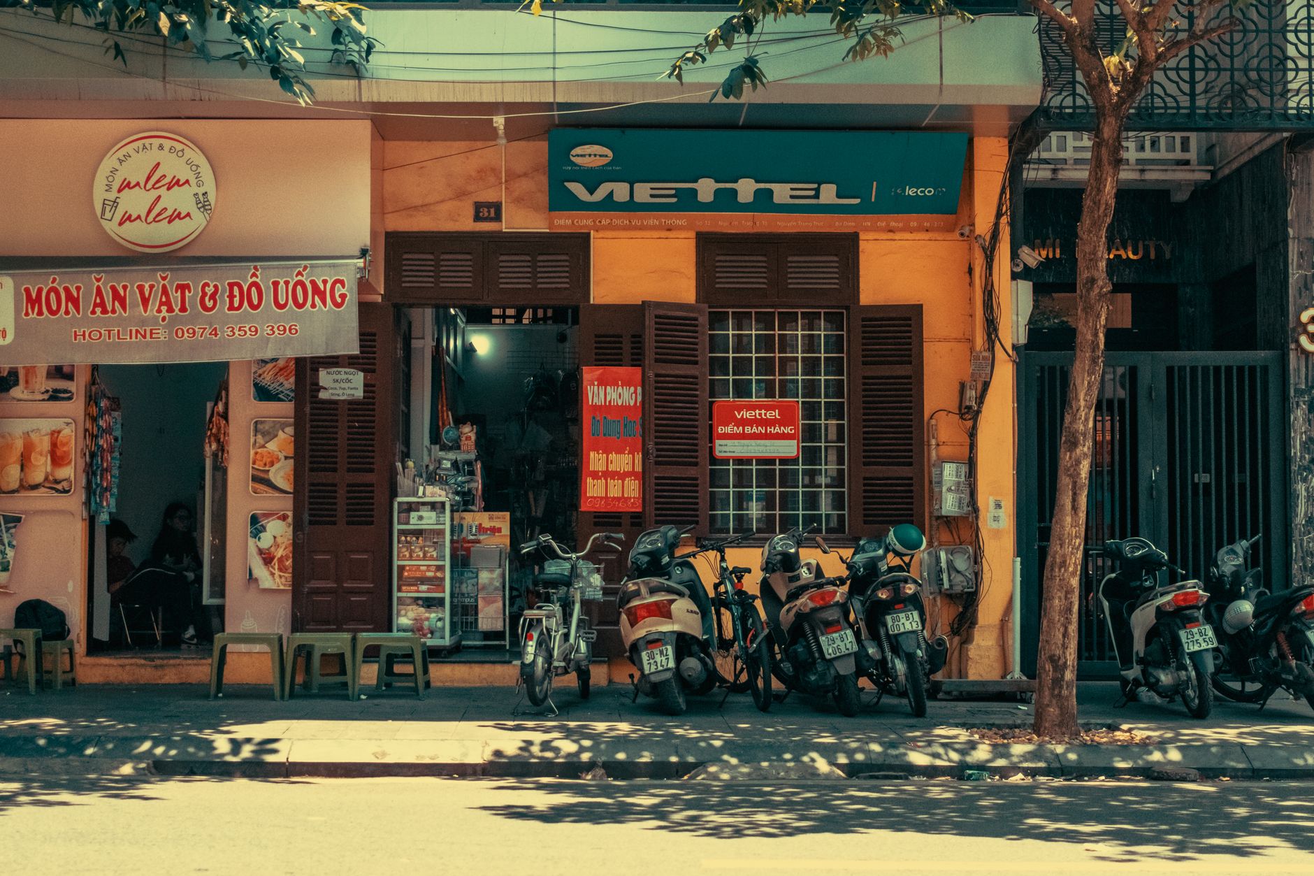Colorful urban street scene with parked scooters, shops, and tree shadows in daylight.