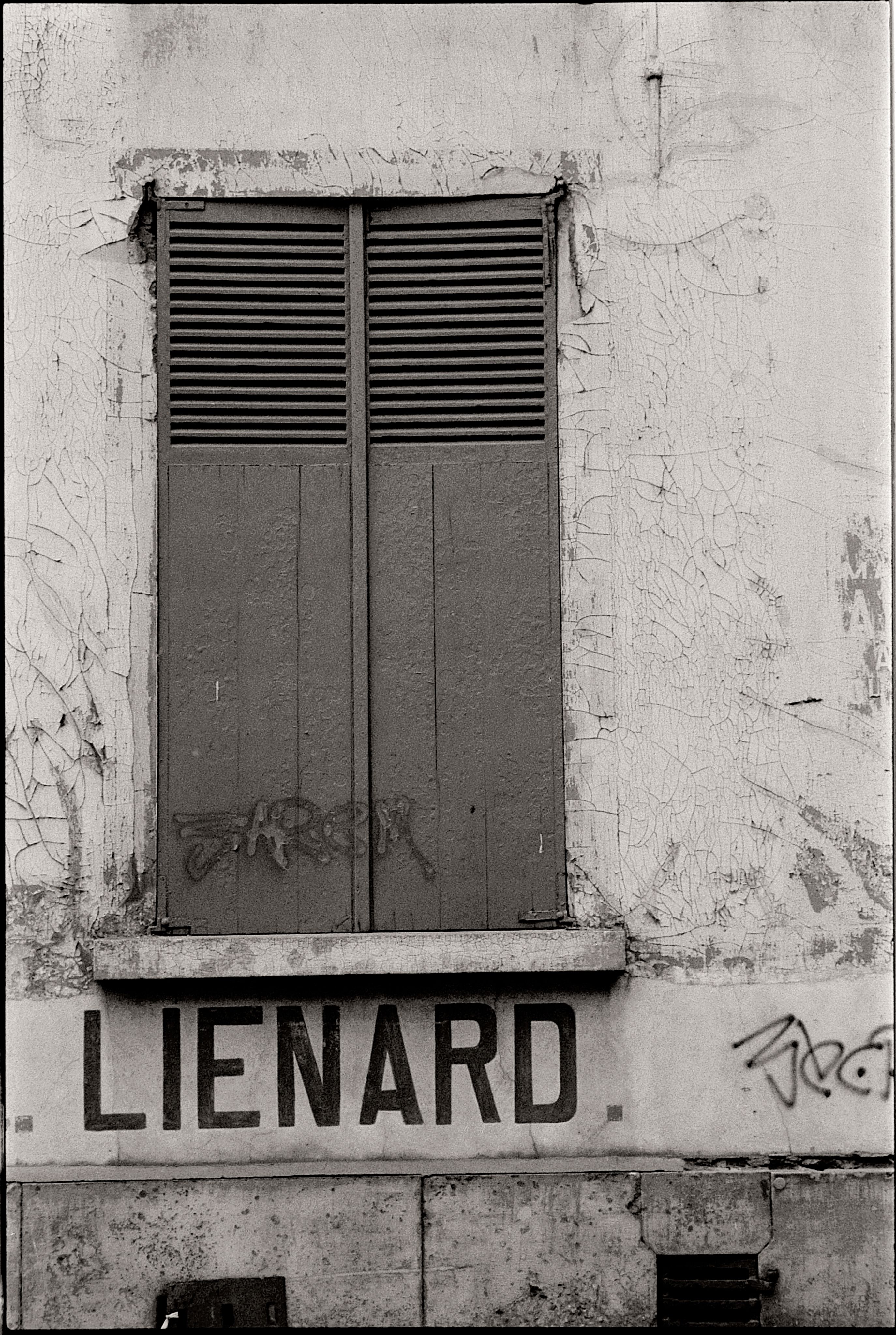 Black and white image of a weathered wall with closed shutters and graffiti, urban setting.