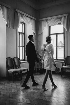 Black and white photo of a couple in formal attire, holding hands in a classic interior setting.