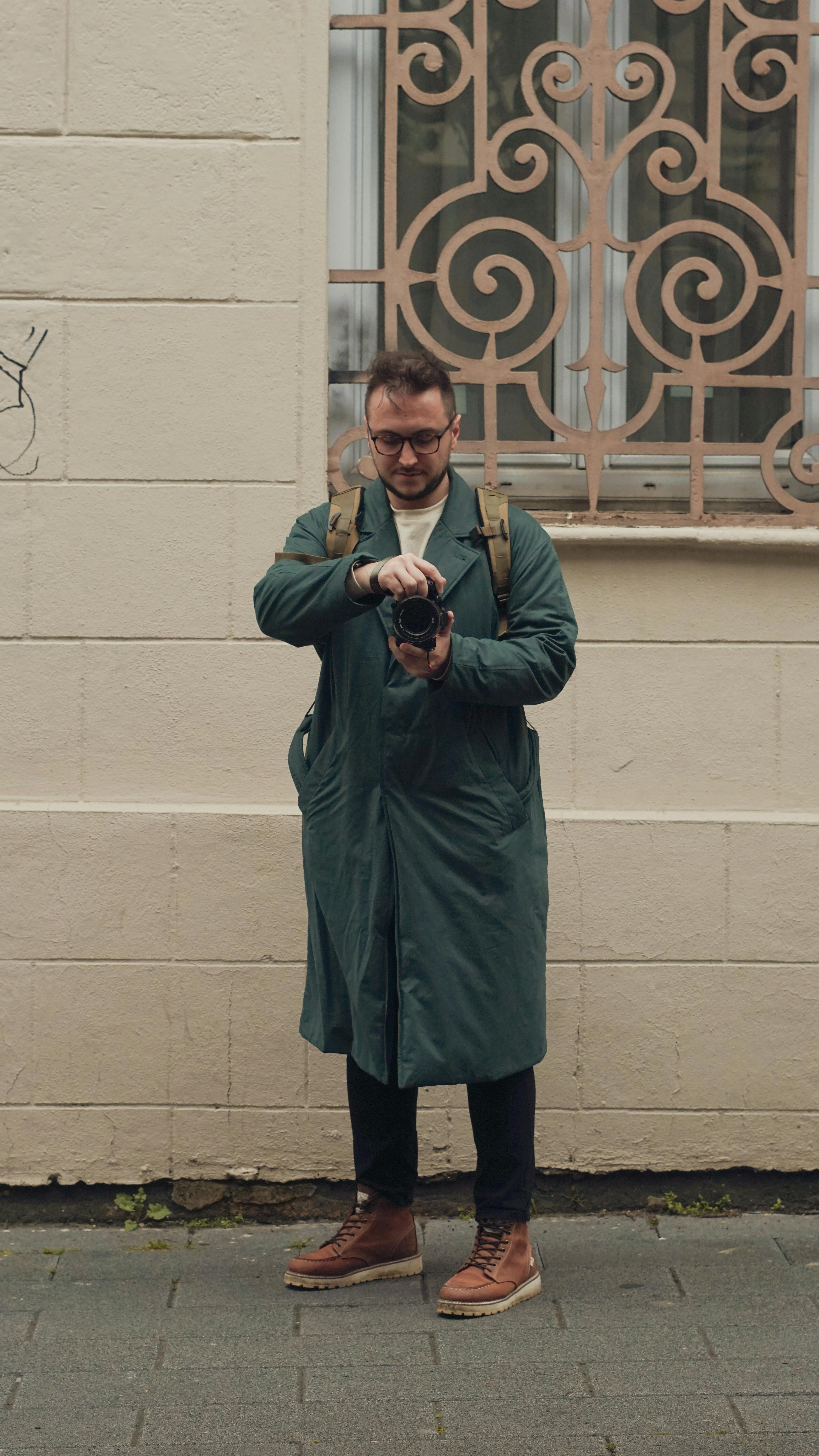 A man in a green coat takes a photo outdoors against a decorative wall. Urban fashion captured.