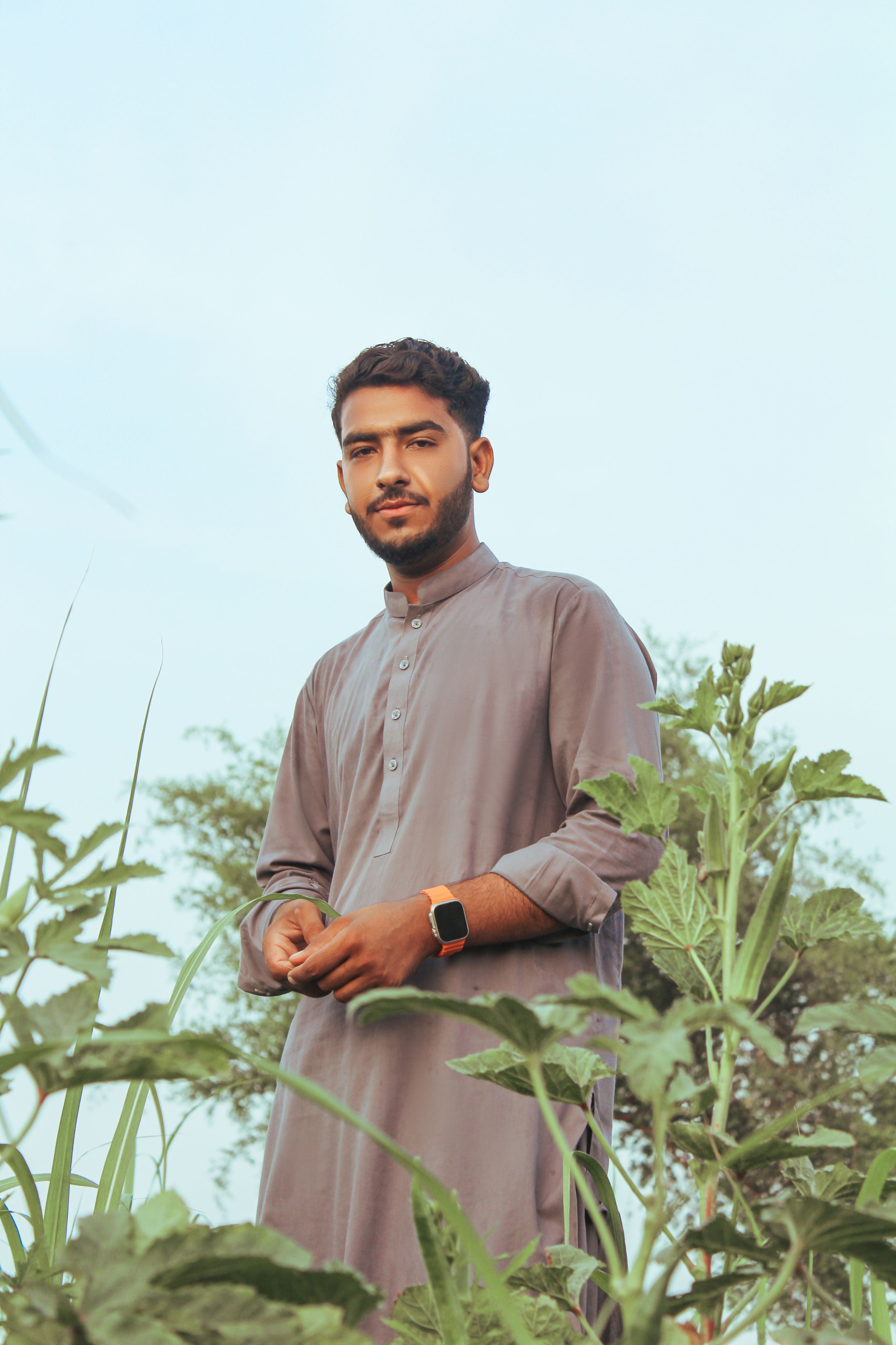 A man in traditional clothing standing amidst greenery with a clear blue sky.