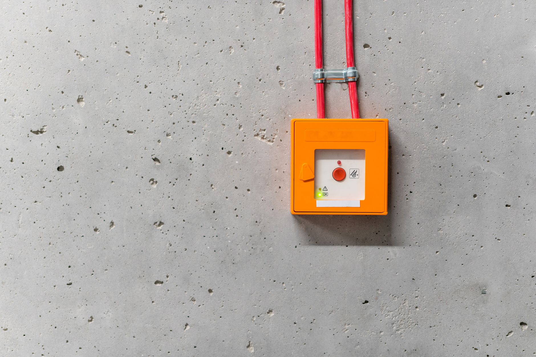 Close-up of an orange emergency stop button with red wires against a concrete wall, indoors.