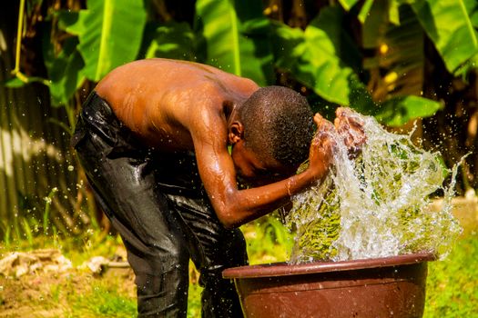 A child washes his head with fresh water from a brown pot in a sunny outdoor setting.