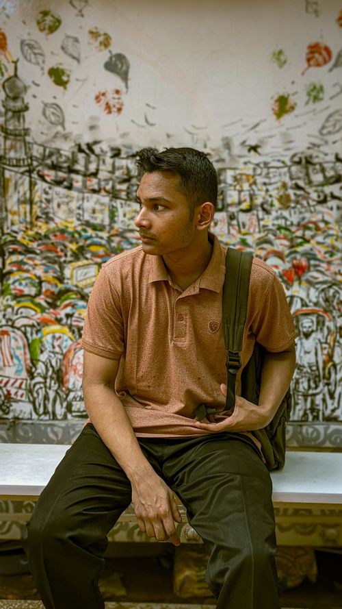 Free Young man in Dhaka seated on a bench, set against a colorful mural backdrop, holding a backpack. Stock Photo