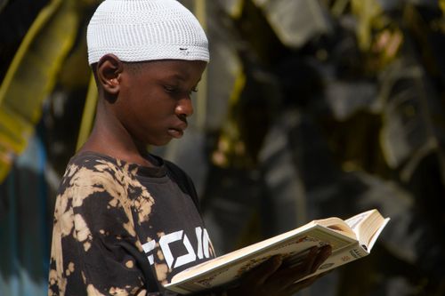 Free Young boy with kufi reading book outdoors, focused and pensive, highlighting cultural and educational themes. Stock Photo