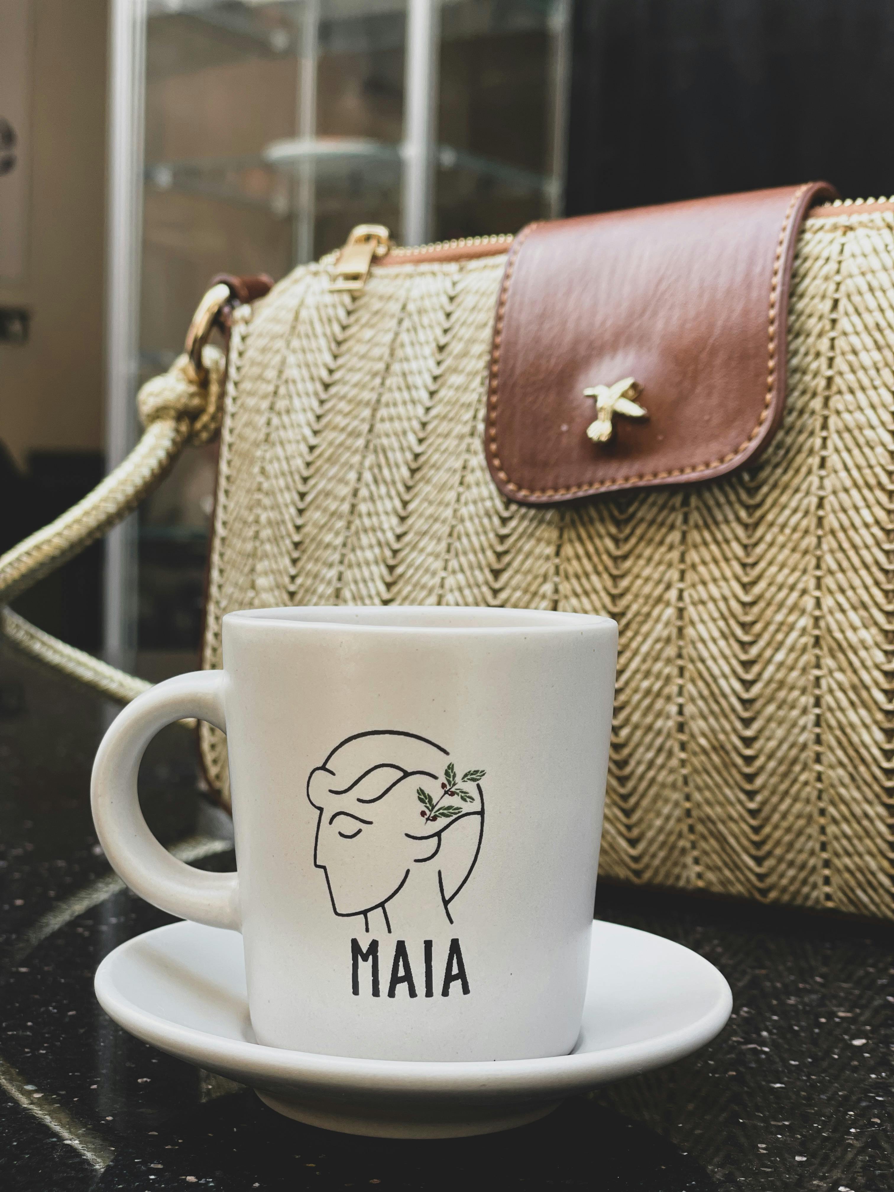 Close-up of a coffee cup with artistic Maia design alongside a stylish handbag on a café table.