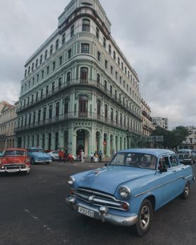 Classic cars drive past a historic building in Havana, Cuba, showcasing vintage architecture and urban life.