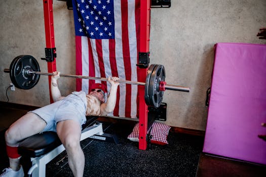 Man bench pressing weights in gym, emphasizing strength and fitness. USA flag in backdrop adds patriotic theme.
