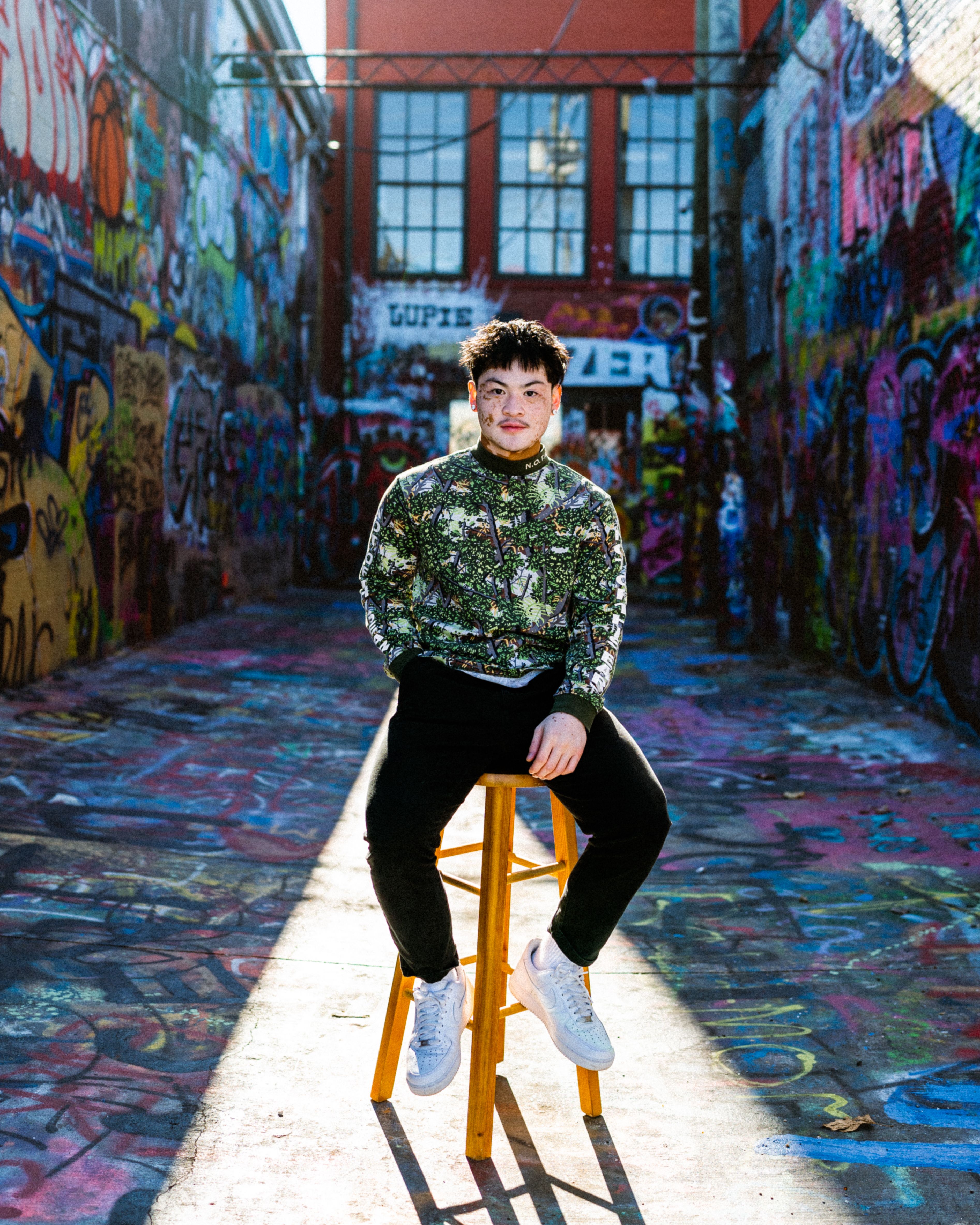 A young man sitting confidently on a stool in a vibrant graffiti-covered alley.