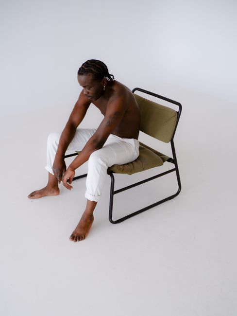 Contemplative topless man seated on a minimalist chair in a bright studio.