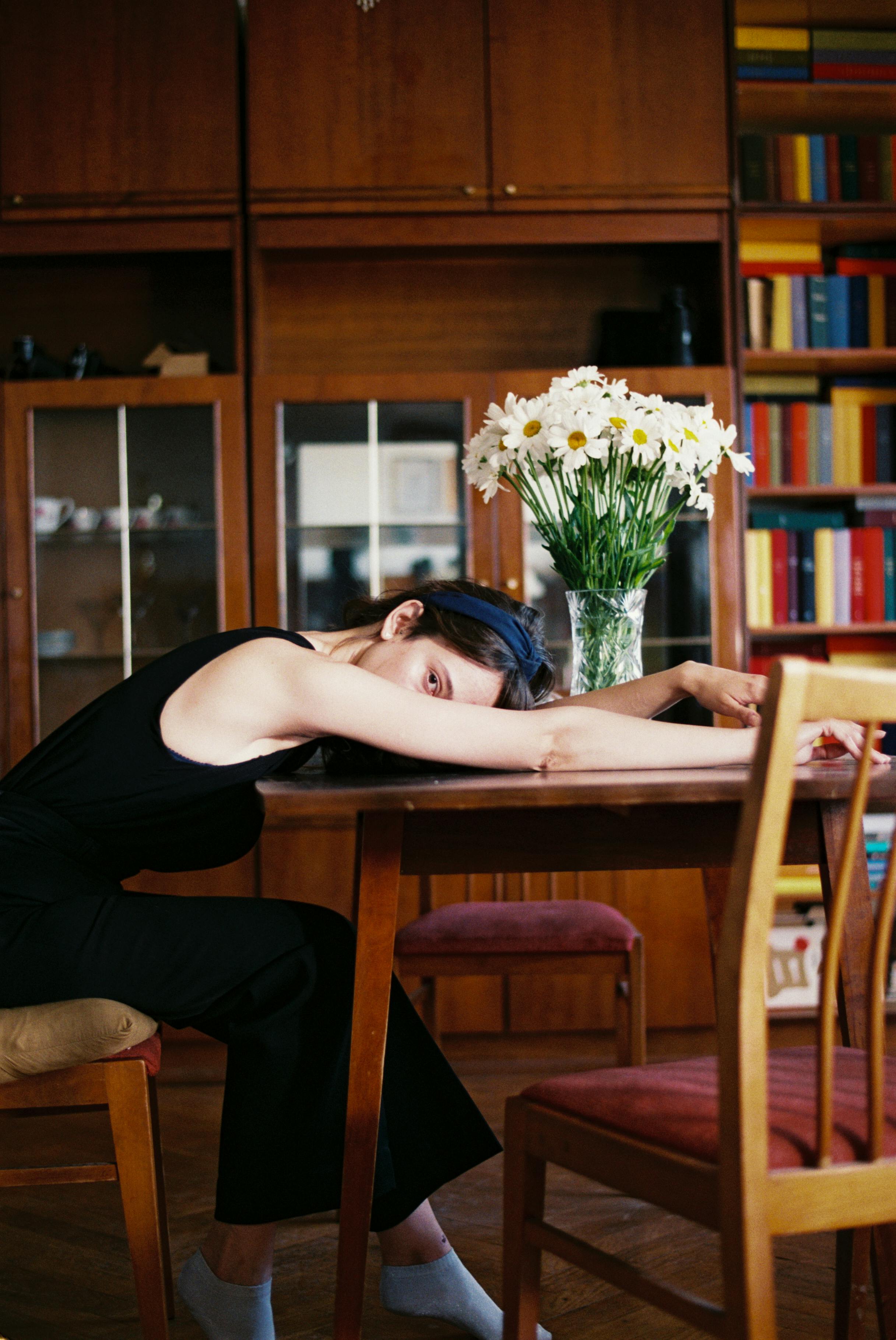 A woman leans over a table in a cozy library, surrounded by flowers and bookshelves.