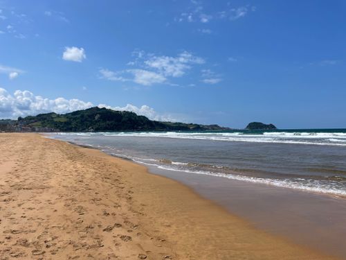 Free Calm sandy beach with waves under a blue sky, featuring green hills in the background. Stock Photo