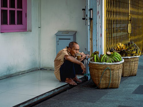 Man Crouching on Ground Next to Baskets of Bananas Sipping Coffee