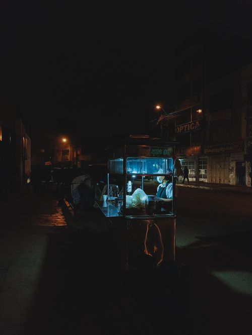 Free A small food stand illuminates a dark urban street at night, creating a moody and dramatic atmosphere. Stock Photo