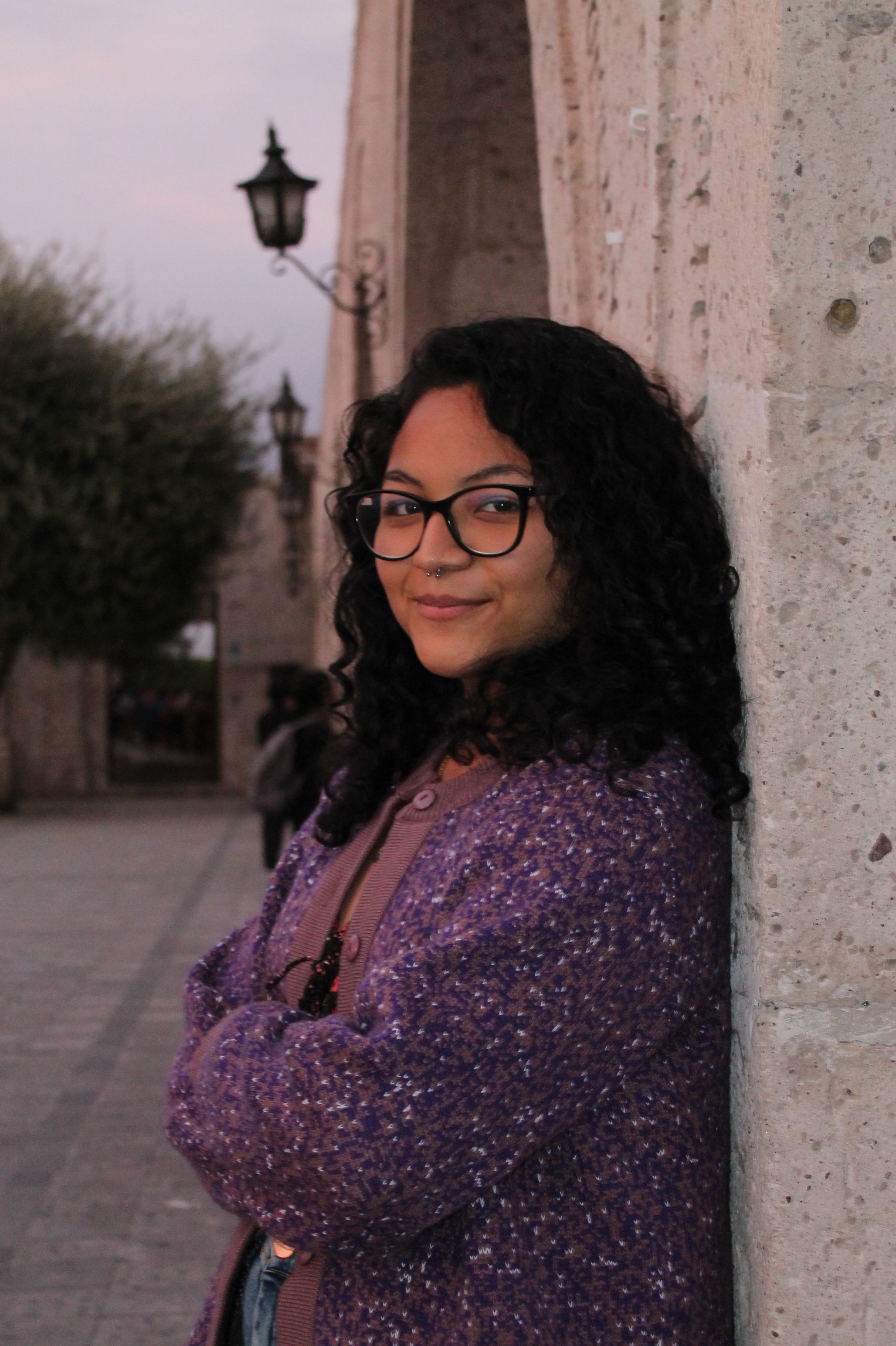 Smiling woman wearing glasses and a purple sweater, leaning against a stone wall outdoors.