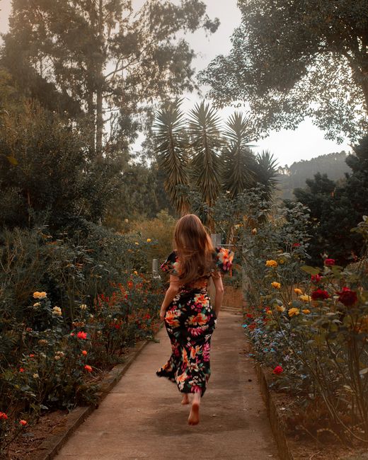 Woman in floral dress walking barefoot along a lush garden path in Brazil, surrounded by colorful flowers.