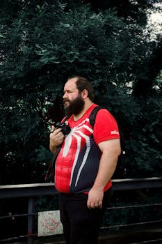 A bearded man stands outdoors with a camera, wearing a Spiderman-themed t-shirt, ready to capture moments.