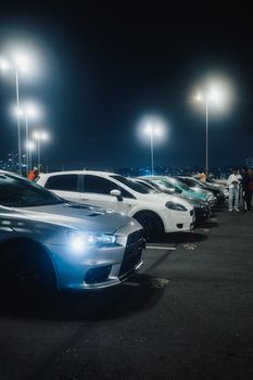 A vivid night scene capturing a row of cars parked under bright streetlights in a spacious parking lot.